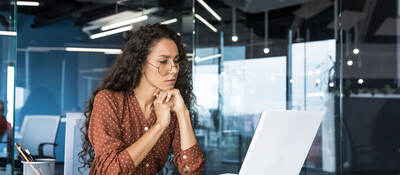 Person in a glass office, looking thoughtfully at a laptop