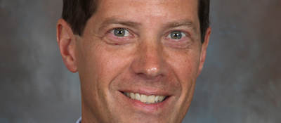 Headshot of Todd Denison wearing a black suit, blue shirt and navy blue tie on a grey background